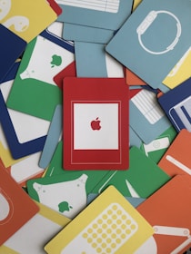 A collection of colorful cards, each showcasing various Apple product silhouettes. The cards are arranged in an overlapping, scattered manner. The central card is red with a white frame and features a prominent red Apple logo. Other cards display images of products like watches, smartphones, and computers in colors like blue, green, yellow, and orange.