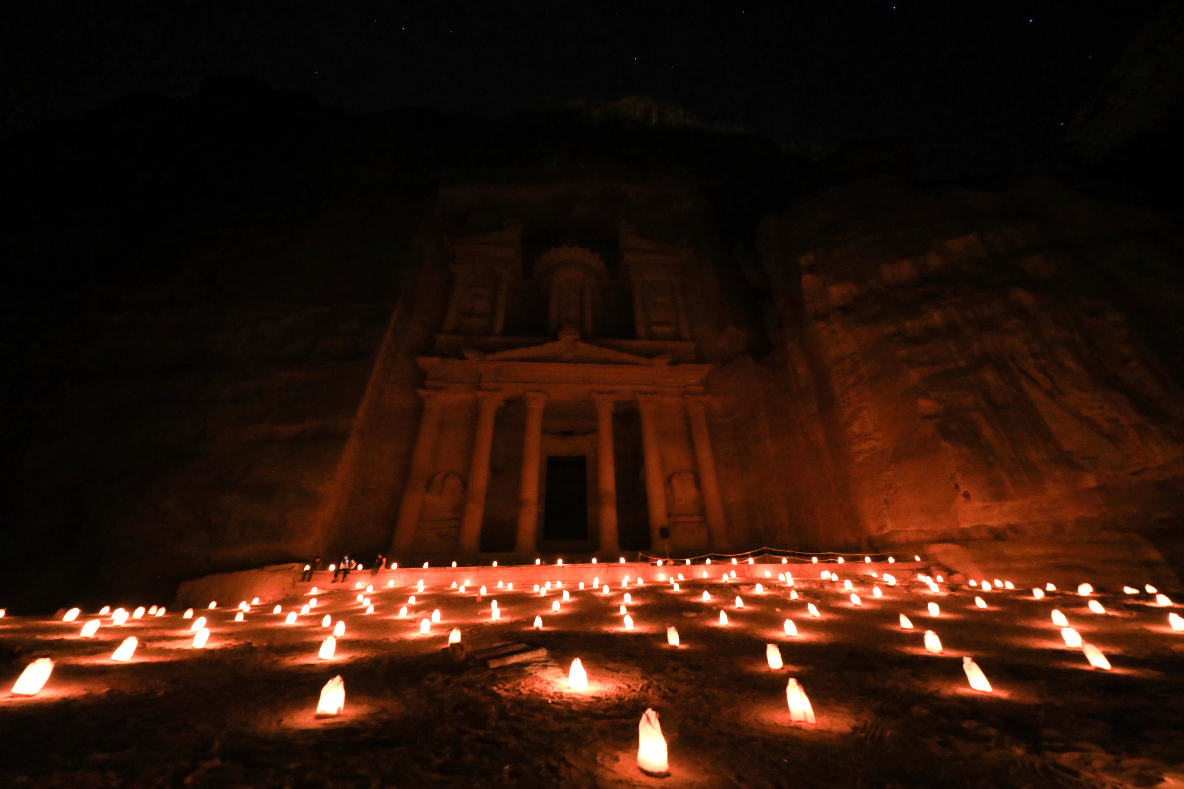 Petra by Night: the Treasury facade glowing amber out of the darkness above a forecourt covered with hundreds of paper-bag candles, their light spilling warm across the sand and up the gorge walls under a starry sky.