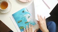 A person is flipping through a magazine page showing a tropical island scene with turquoise water and lush greenery. A teacup with amber liquid is placed on a saucer to the side. The hands have neatly painted nails, and a pen and a pink plate are also visible on the table.