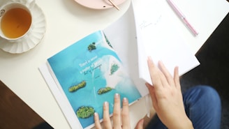 A person is flipping through a magazine page showing a tropical island scene with turquoise water and lush greenery. A teacup with amber liquid is placed on a saucer to the side. The hands have neatly painted nails, and a pen and a pink plate are also visible on the table.