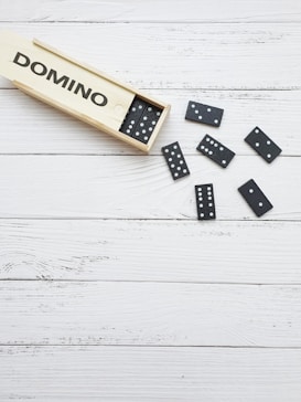 A wooden box containing black domino tiles rests on a white wooden surface. Several domino pieces are scattered around the box, each with a pattern of white dots.