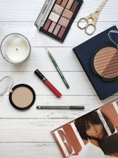 A flat lay image featuring a variety of beauty and makeup items arranged on a wooden surface. Items include an eyeshadow palette with multiple shades, a red lipstick, a green eyeliner pencil, face powders in circular containers, a pair of golden scissors, and a fashion magazine. A white candle is also present, adding a touch of elegance to the arrangement.
