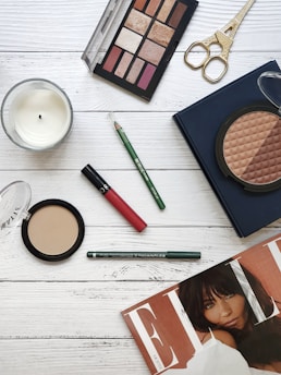 A flat lay image featuring a variety of beauty and makeup items arranged on a wooden surface. Items include an eyeshadow palette with multiple shades, a red lipstick, a green eyeliner pencil, face powders in circular containers, a pair of golden scissors, and a fashion magazine. A white candle is also present, adding a touch of elegance to the arrangement.