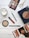A flat lay image featuring a variety of beauty and makeup items arranged on a wooden surface. Items include an eyeshadow palette with multiple shades, a red lipstick, a green eyeliner pencil, face powders in circular containers, a pair of golden scissors, and a fashion magazine. A white candle is also present, adding a touch of elegance to the arrangement.