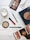 A flat lay image featuring a variety of beauty and makeup items arranged on a wooden surface. Items include an eyeshadow palette with multiple shades, a red lipstick, a green eyeliner pencil, face powders in circular containers, a pair of golden scissors, and a fashion magazine. A white candle is also present, adding a touch of elegance to the arrangement.