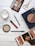 A flat lay image featuring a variety of beauty and makeup items arranged on a wooden surface. Items include an eyeshadow palette with multiple shades, a red lipstick, a green eyeliner pencil, face powders in circular containers, a pair of golden scissors, and a fashion magazine. A white candle is also present, adding a touch of elegance to the arrangement.