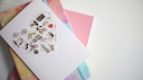 A greeting card with a heart-shaped collage featuring miniature icons and text, possibly themed around travel or city life. It is placed on top of a stack of colorful notebooks with abstract patterns in pastel shades.