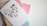 A greeting card with a heart-shaped collage featuring miniature icons and text, possibly themed around travel or city life. It is placed on top of a stack of colorful notebooks with abstract patterns in pastel shades.