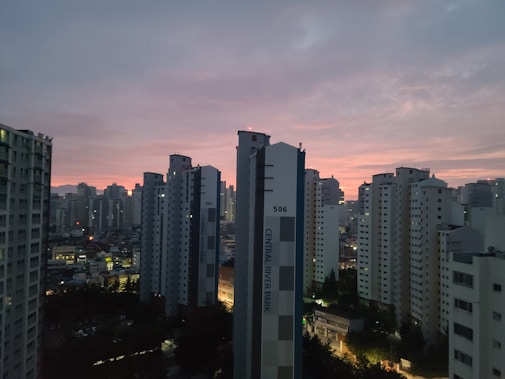 High-rise apartment buildings are silhouetted against a sunset sky with hues of pink and orange. The cityscape features densely packed skyscrapers, reflecting urban life. There are a few scattered lights from windows and streetlights, indicating early evening.
