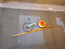 A gray wall features a bicycle mounted as a decorative piece. The rear wheel of the bicycle is replaced with a large, colorful clock face, predominantly orange and yellow. A wooden beam intersects the bicycle at an angle, adding a dynamic element to the composition. The background includes a roughly textured section of white brick at the top.