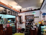 A cozy cafe interior with wooden counters and decorative shelves. There are various action figures and trinkets displayed above the counter. A staff member is preparing drinks behind the counter, surrounded by coffee-making equipment. Menus with Korean writing are visible, featuring drink options. The ambiance is warm and eclectic, with touches of vintage decor.