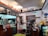 A cozy cafe interior with wooden counters and decorative shelves. There are various action figures and trinkets displayed above the counter. A staff member is preparing drinks behind the counter, surrounded by coffee-making equipment. Menus with Korean writing are visible, featuring drink options. The ambiance is warm and eclectic, with touches of vintage decor.