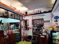 A cozy cafe interior with wooden counters and decorative shelves. There are various action figures and trinkets displayed above the counter. A staff member is preparing drinks behind the counter, surrounded by coffee-making equipment. Menus with Korean writing are visible, featuring drink options. The ambiance is warm and eclectic, with touches of vintage decor.