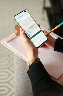 A person holding a smartphone with both hands. The phone displays a social media app with images and text. The person has red-painted nails and wears a black long-sleeve top. A lined notebook rests on their lap, with a copper clip holding the pages. A pencil with a blue tip is also visible.