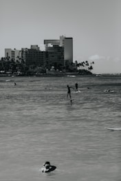 Several people are engaging in water activities such as paddleboarding and swimming in front of a backdrop of tall apartment buildings and palm trees along the shoreline.