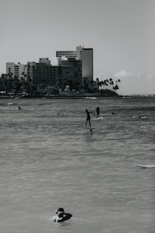 Several people are engaging in water activities such as paddleboarding and swimming in front of a backdrop of tall apartment buildings and palm trees along the shoreline.