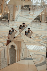 A complex geometric mirror structure reflects multiple angles of an intricate interior with ornate decorations and patterns. Two individuals, wearing masks, pose with cameras, capturing different perspectives within the reflections. Their images are repeated across the fragmented mirror surfaces. In the background, exhibition stands with framed artworks can be seen, and a person is walking through the space.