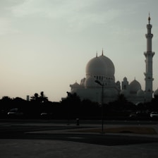 A majestic mosque with large domes and a prominent minaret is silhouetted against a cloudy sky. The structure appears grand and serene, set against a backdrop of palm trees and an empty paved plaza.