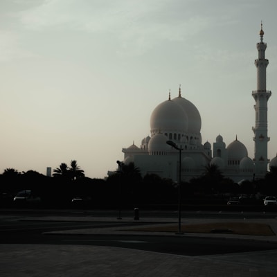 A majestic mosque with large domes and a prominent minaret is silhouetted against a cloudy sky. The structure appears grand and serene, set against a backdrop of palm trees and an empty paved plaza.