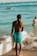 A person with braided hair is standing on a sandy beach facing the ocean, holding a surfboard. They are wearing bright turquoise shorts with tropical patterns. In the background, other people are swimming and enjoying the water under a clear blue sky.