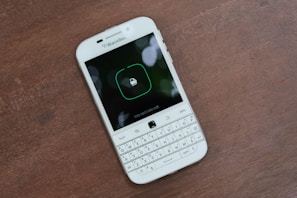 A white BlackBerry smartphone is placed on a wooden surface. The phone has a physical QWERTY keyboard and its screen displays a lock icon with Vietnamese text underneath.