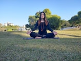 A person is meditating outdoors on a grass field, seated in a cross-legged position with eyes closed. They are wearing black attire and a beaded necklace, sitting on a mat. The background includes trees, greenery, and some buildings under a clear blue sky.