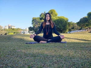 A person is meditating outdoors on a grass field, seated in a cross-legged position with eyes closed. They are wearing black attire and a beaded necklace, sitting on a mat. The background includes trees, greenery, and some buildings under a clear blue sky.