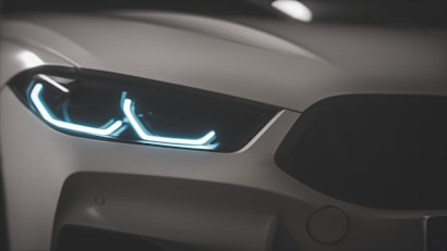 A close-up view of a car's front headlight with bright, futuristic LED lights. The design is sleek and modern, emphasizing the sharp contours and metallic sheen of the vehicle.