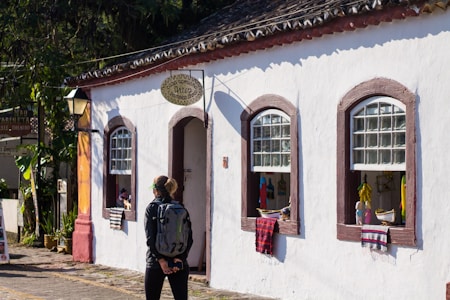 A person with a backpack stands in front of a quaint, whitewashed building with decorative arched windows. The windows display several colorful handcrafted items, including small figures and textiles. The building has a rustic sign, and a cobblestone street runs alongside it.