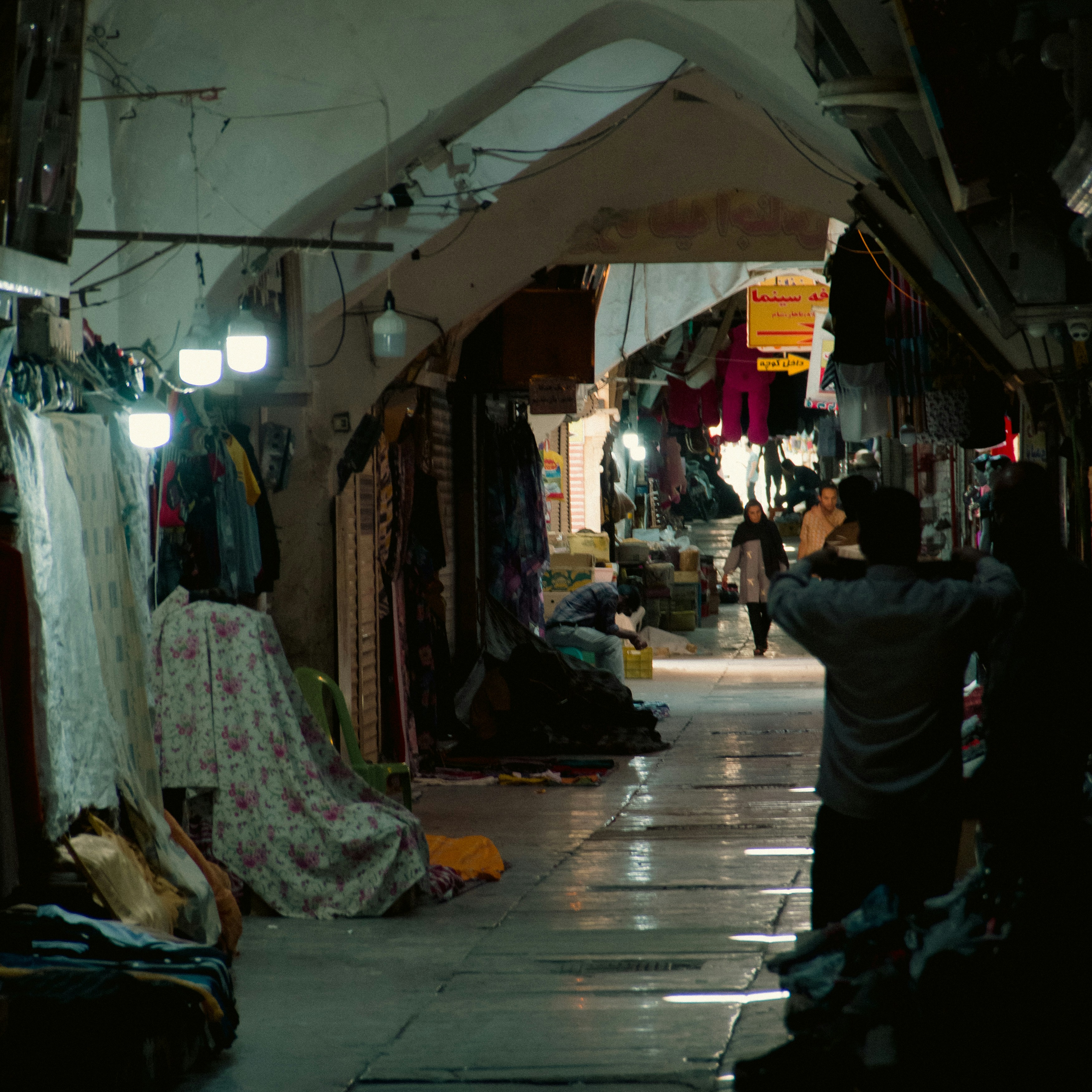 Busy marketplace with diverse goods displayed on tables under warm indoor lighting, dark wooden surfaces, shallow depth of field