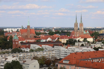 A scenic view of a European cityscape featuring a mix of historical and modern architecture. Prominent gothic churches with tall spires are surrounded by red-roofed buildings and lush greenery. Residential buildings with a modern design appear in the foreground.