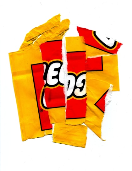 Torn pieces of yellow and red packaging are scattered, with part of a logo visible. The fragments are irregularly shaped and arranged randomly against a white background.