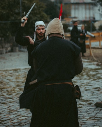 Two people are dressed in medieval-style clothing, engaging in what appears to be a mock combat or reenactment. One person is holding a sword raised above their head as if preparing to strike. The scene takes place outdoors on a cobblestone surface, with a few people observing in the background.