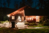 A cozy wooden house is adorned with string lights, creating a warm and inviting ambiance. The patio features outdoor furniture including a grill and chairs, surrounded by lush greenery. Soft, warm light emanates from the windows, suggesting a comforting interior setting.