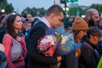 A group of people, gathered closely together outdoors, appear to be in a moment of silence or contemplation. One person in the foreground holds a bouquet of flowers. The mood suggests a somber or reflective atmosphere.