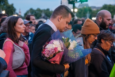 A group of people, gathered closely together outdoors, appear to be in a moment of silence or contemplation. One person in the foreground holds a bouquet of flowers. The mood suggests a somber or reflective atmosphere.