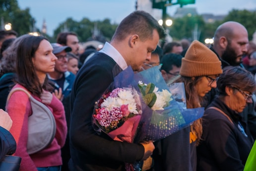 A group of people, gathered closely together outdoors, appear to be in a moment of silence or contemplation. One person in the foreground holds a bouquet of flowers. The mood suggests a somber or reflective atmosphere.