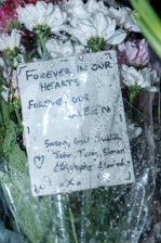 A bouquet of white and pink flowers wrapped in cellophane, with raindrops on the surface. A handwritten card with a heartfelt message is attached, expressing remembrance.