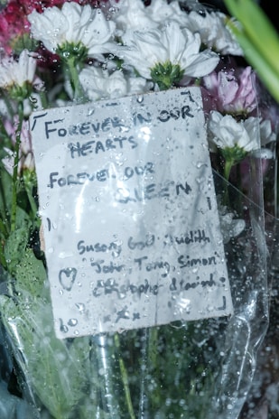 A bouquet of white and pink flowers wrapped in cellophane, with raindrops on the surface. A handwritten card with a heartfelt message is attached, expressing remembrance.