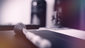 A close-up of a makeup brush resting on a smooth surface with a blurred background, suggesting a focus on the bristles and handle. The lighting is soft, creating a calm and serene atmosphere with touches of light reflections.