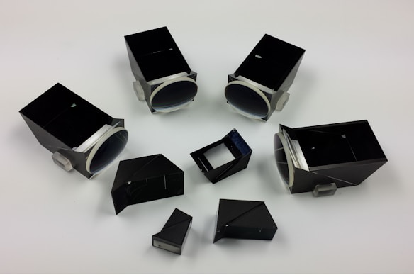 A variety of black optical devices and prism-like components are arranged on a plain white background. The devices appear to be made of a combination of black material and transparent elements, with some featuring cylindrical, lens-like ends.
