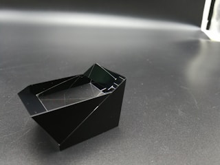 A geometric, angular black object rests on a dark surface with a slightly reflective quality, suggesting a matte material. The object appears to have sharp, defined edges and a hollow interior, creating intriguing light reflections and shadows.