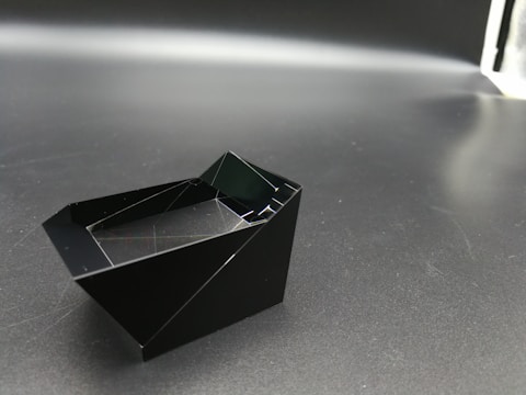 A geometric, angular black object rests on a dark surface with a slightly reflective quality, suggesting a matte material. The object appears to have sharp, defined edges and a hollow interior, creating intriguing light reflections and shadows.