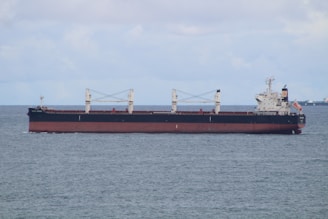 A large cargo ship is sailing on a calm sea under a partly cloudy sky. The vessel is predominantly black with a red hull and has white superstructures with cranes and a flag at the stern.
