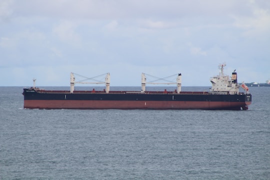 A large cargo ship is sailing on a calm sea under a partly cloudy sky. The vessel is predominantly black with a red hull and has white superstructures with cranes and a flag at the stern.