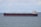 A large cargo ship is sailing on a calm sea under a partly cloudy sky. The vessel is predominantly black with a red hull and has white superstructures with cranes and a flag at the stern.