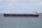 A large cargo ship is sailing on a calm sea under a partly cloudy sky. The vessel is predominantly black with a red hull and has white superstructures with cranes and a flag at the stern.