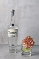 A bottle of Tanteo Blanco tequila is positioned next to a glass with a slice of lime and a peach-colored carnation inside. The setting is minimalist with a light concrete background and surface, creating a clean and sophisticated presentation.