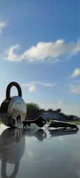 A padlock and a set of keys are placed on a reflective surface with a clear blue sky and some white clouds in the background. The padlock is metallic with a visible brand name, and the keys are lying next to it, casting a shadow.