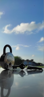 A padlock and a set of keys are placed on a reflective surface with a clear blue sky and some white clouds in the background. The padlock is metallic with a visible brand name, and the keys are lying next to it, casting a shadow.